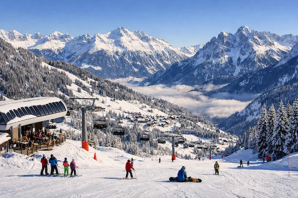 Skifahrer auf einer sonnigen Skipiste in den Alpen mit Seilbahn, Bergrestaurant und schneebedecktem Bergpanorama im Hintergrund.