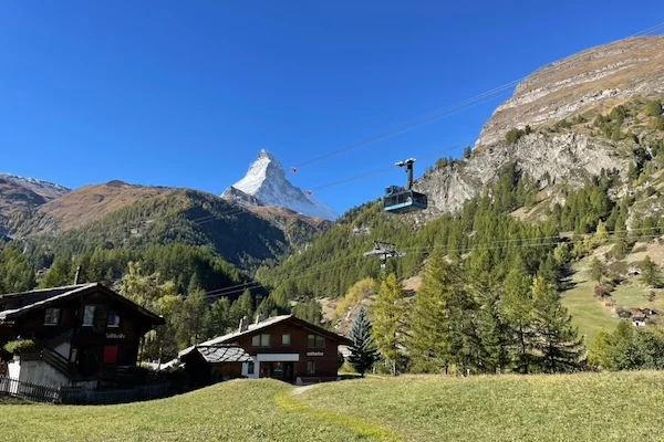 Alpenlandschaft in der Schweiz mit Blick auf das Matterhorn, Seilbahn über grünen Wiesen, traditionelle Chalets und bewaldete Berghänge – ideales Ziel für Tagesausflüge.