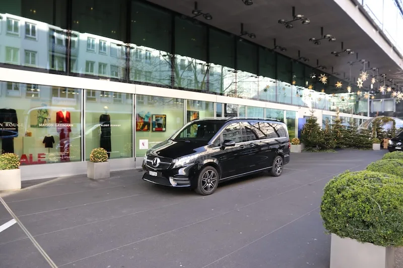A black Mercedes-Benz van from a business chauffeur service in front of a modern building in Zurich, prepared for an exclusive transfer.