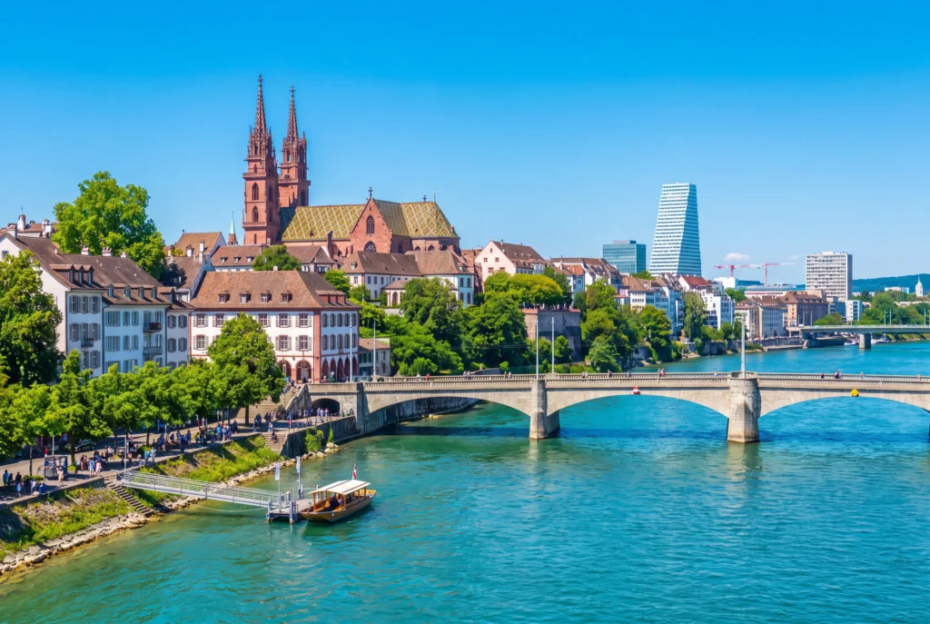 Basel Ausflug ab Zürich mit Chauffeur – das rote Basler Münster aus Sandstein mit seinen Doppeltürmen und dem bunt gemusterten Ziegeldach über der Altstadt, die Mittlere Brücke über den türkisblauen Rhein, eine hölzerne Fähre und der markante Roche-Turm in der modernen Skyline