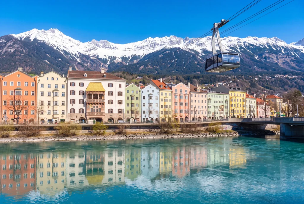 Innsbruck Privattour ab Zürich mit Chauffeur – die berühmte Reihe bunter Pastellhäuser am Mariahilf-Ufer spiegelt sich im türkisgrünen Inn, darüber schwebt eine Gondel der Nordkettenbahn vor der schneebedeckten Bergkette der Nordkette unter blauem Himmel