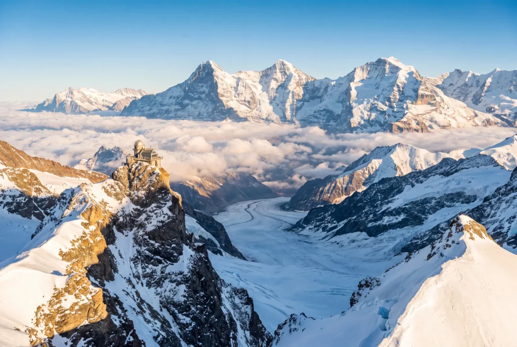 Jungfraujoch Ausflug ab Zürich mit Chauffeur – das Sphinx-Observatorium auf dem Felsgipfel über dem geschwungenen Aletschgletscher, die schneebedeckten Gipfel von Eiger, Mönch und Jungfrau über einem Wolkenmeer im goldenen Abendlicht