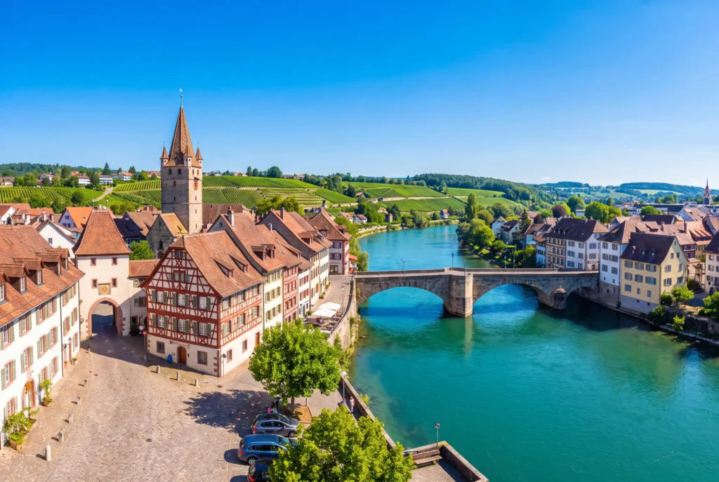 Rheinfelden ab Zürich mit Chauffeur – die mittelalterliche Altstadt mit Torturm, Fachwerkhäusern und gepflastertem Platz am Ufer des türkisblauen Rheins, verbunden durch die alte steinerne Bogenbrücke, dahinter sanfte Weinberghänge unter klarem blauem Himmel