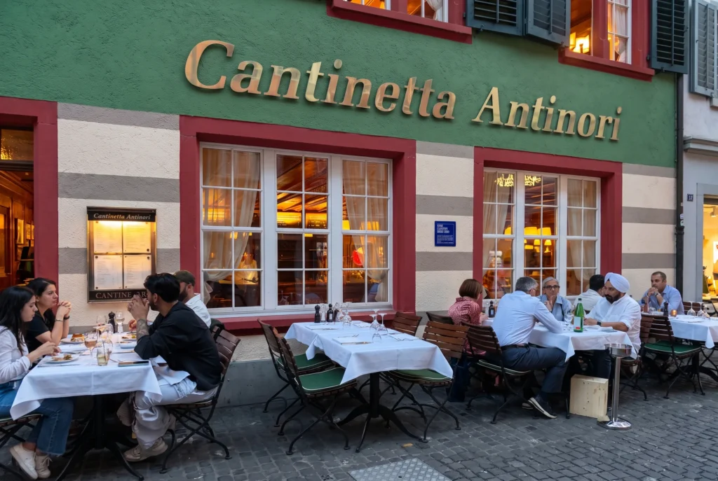 Cantinetta Antinori ab Zürich mit Chauffeur – die markante grün-weiss-rot gestreifte Fassade des italienischen Restaurants in der Zürcher Altstadt mit goldenem Schriftzug, Gästen an weiss eingedeckten Tischen auf der Kopfsteinpflaster-Terrasse im warmen Abendlicht