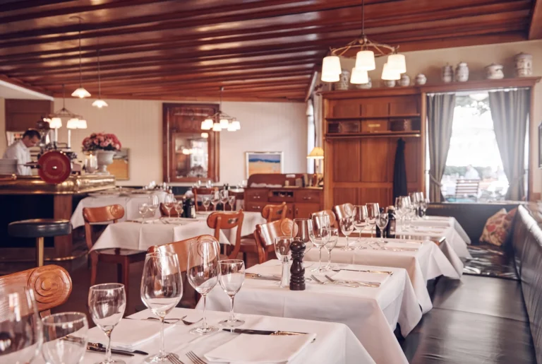Cantinetta Antinori ab Zürich mit Chauffeur – der warme holzvertäfelte Speisesaal mit Kassettendecke, Pendelleuchten, weiss eingedeckten Tischen mit Weingläsern, einer Aufschnittmaschine an der Theke und Blick zur Terrasse