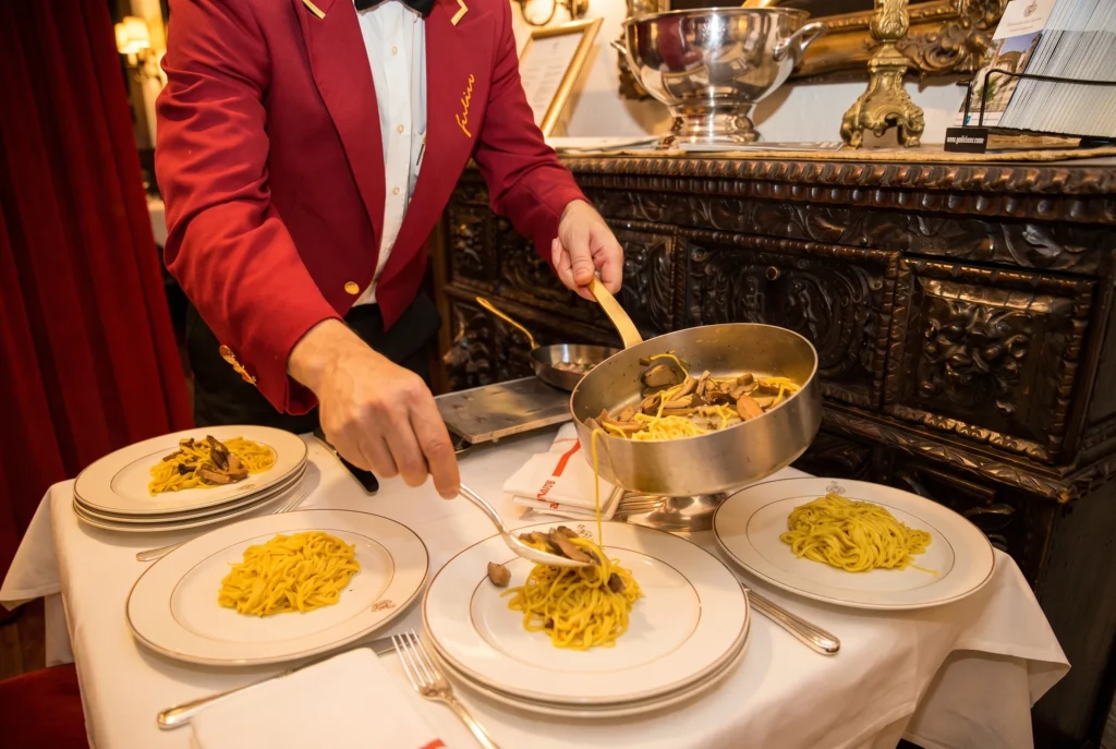 Casa Ferlin ab Zürich mit Chauffeur – ein Kellner im roten Jackett mit goldener Ferlin-Stickerei serviert am Tisch frische Tagliolini mit Steinpilzen aus einer Kupferpfanne auf Teller mit goldenem Wappen, im traditionsreichen italienischen Restaurant