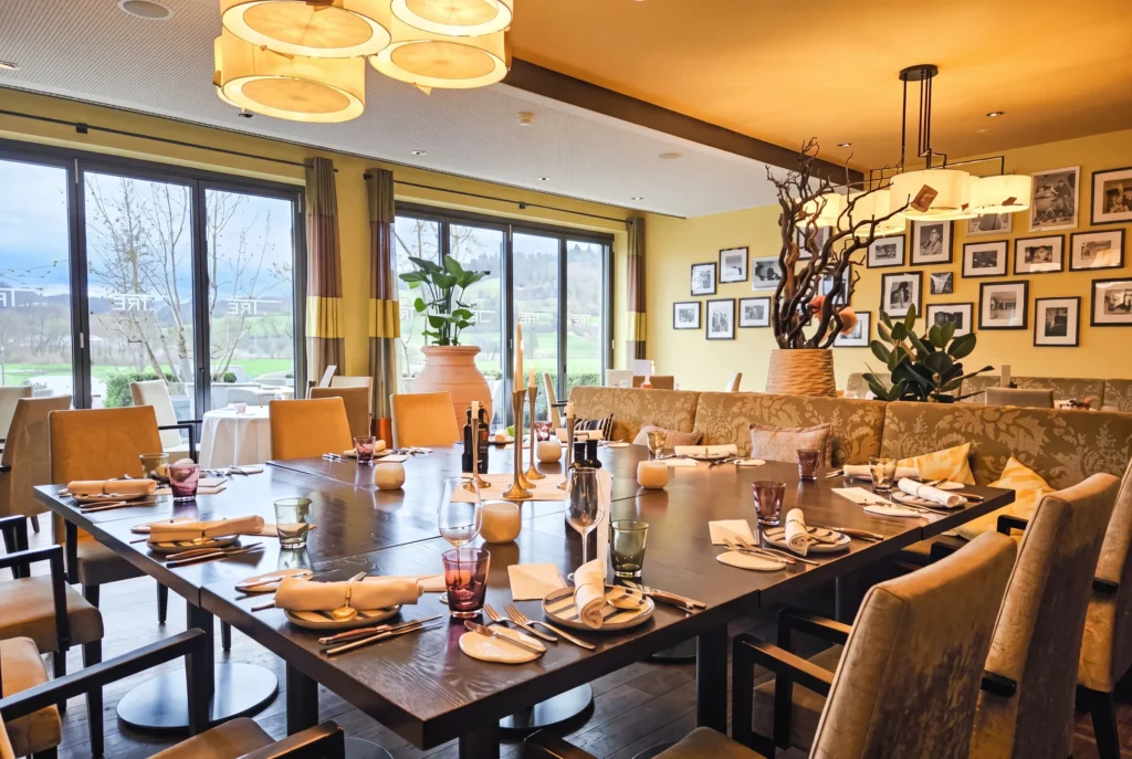 Osteria Tre ab Zürich mit Chauffeur – der elegante Speisesaal des italienischen Restaurants mit gelben Wänden, einer schwarz-weissen Fotogalerie, eingedeckten dunklen Holztischen mit farbigen Gläsern und Blick durch grosse Fenster ins grüne Umland