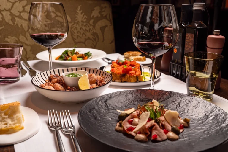 Osteria Tre ab Zürich mit Chauffeur – italienische Gerichte auf gedecktem Tisch mit zwei Gläsern Rotwein, frittierten Calamari mit Zitrone, Tomaten-Bruschetta und einem fein angerichteten Vitello-Teller mit Granatapfelkernen und Kapern