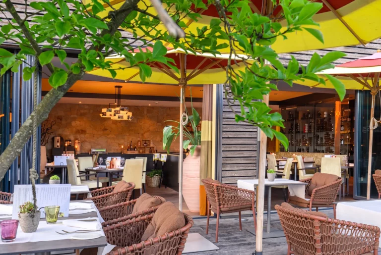 Osteria Tre ab Zürich mit Chauffeur – die überdachte Terrasse des Restaurants mit gelb-roten Sonnenschirmen, geflochtenen Rattansesseln und eingedeckten Tischen, im Hintergrund der Innenraum mit Natursteinwand und gut gefülltem Weinregal