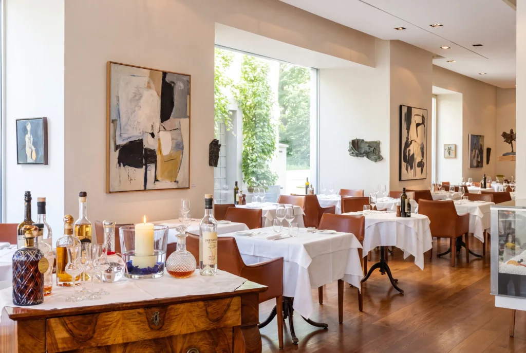 Ristorante Bianchi ab Zürich mit Chauffeur – der helle elegante Speisesaal des Fischrestaurants mit moderner abstrakter Kunst an cremefarbenen Wänden, cognacfarbenen Lederstühlen an weiss eingedeckten Tischen, einer antiken Nussbaum-Kommode mit edlen Spirituosen und bodentiefen Fenstern zum grünen Garten