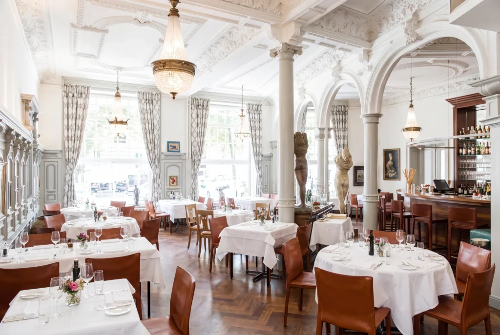 Ristorante Conti ab Zürich mit Chauffeur – der prunkvolle Speisesaal des italienischen Restaurants mit hoher Stuckdecke, funkelnden Kristalllüstern, klassizistischen Säulen, cognacfarbenen Lederstühlen an weiss eingedeckten Tischen und einer langen Bar mit Skulpturen