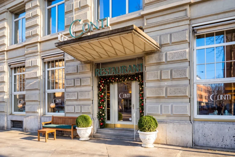 Ristorante Conti ab Zürich mit Chauffeur – der Eingang des Restaurants Conti in einem Sandsteingebäude mit grün-weissem Schriftzug, weihnachtlich geschmücktem Türbogen, Holzbank und kugelförmig geschnittenen Buchsbäumen bei Sonnenschein