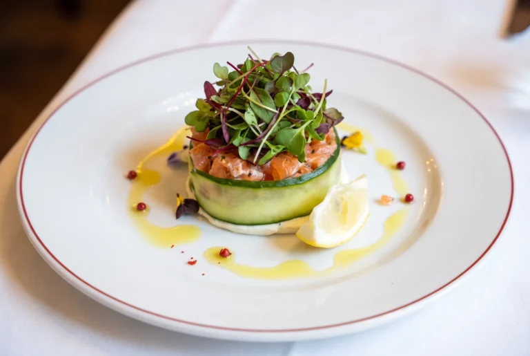 Ristorante Conti ab Zürich mit Chauffeur – ein fein angerichtetes Lachstatar im Gurkenring, gekrönt mit frischen Microgreens, dazu eine Zitronenspalte, rosa Pfefferbeeren und Olivenöl auf einem weissen Teller mit rotem Rand