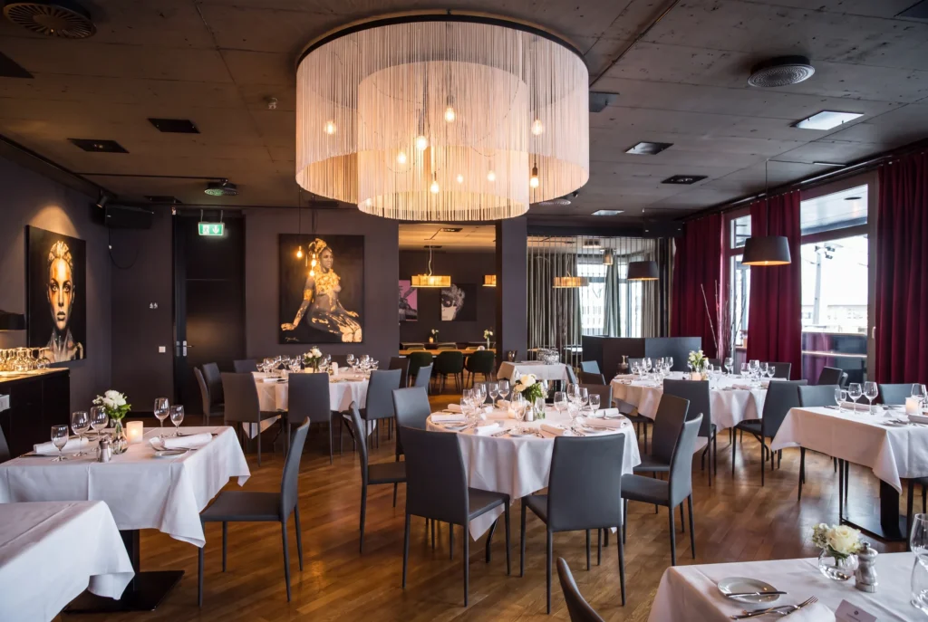 Restaurant Roots in Basel – stimmungsvoller Speisesaal mit grossem Fransen-Kronleuchter, dunklen Wänden, Kunstporträts und bordeauxroten Vorhängen, Fine-Dining Restaurant von Pascal Steffen, Restaurant-Transfer ab Zürich mit Chauffeur