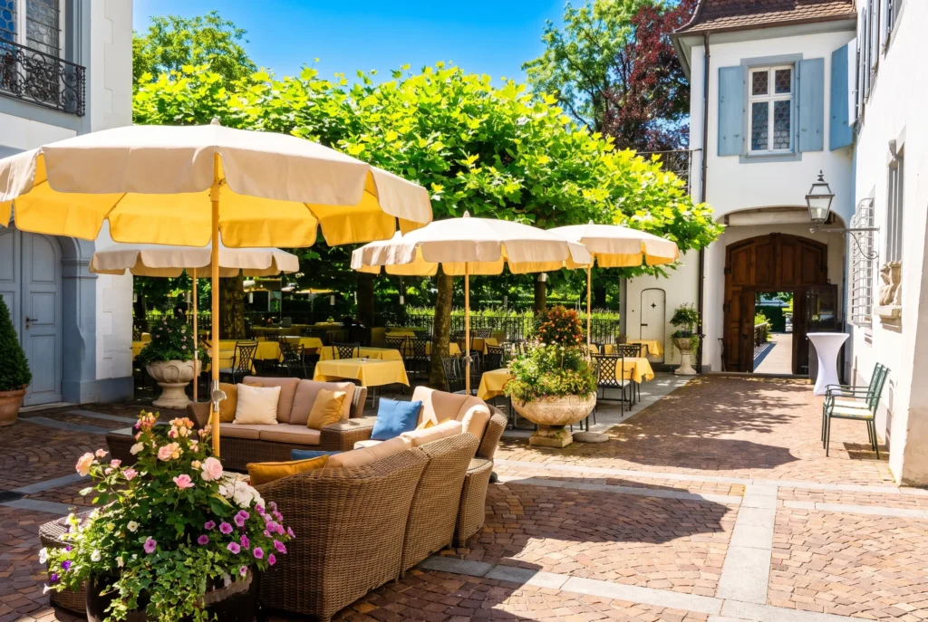 Schloss Bottmingen bei Basel – sonnige Schlossterrasse mit gelben Sonnenschirmen, Lounge-Möbeln und Blumen im historischen Innenhof, Fine-Dining Restaurant im Wasserschloss, Restaurant-Transfer ab Zürich mit Chauffeur