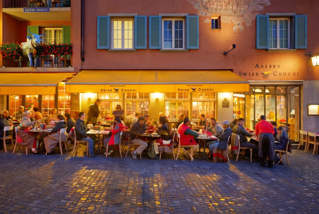 Adler's Swiss Chuchi in der Zürcher Altstadt – beleuchtete Fassade mit gelber Markise und Gästen auf der Terrasse am Kopfsteinpflaster im Niederdorf, Schweizer Spezialitätenrestaurant, Restaurant-Transfer ab Zürich mit Chauffeur